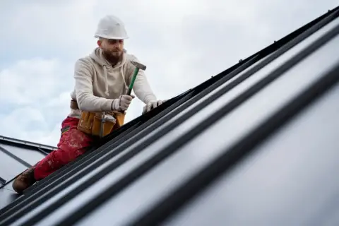 Dekarz w kasku i z młotkiem pracuje na metalowym dachu.