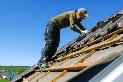 Dekarz w czapce i rękawiczkach pracuje na dachu, naprawiając dachówki.